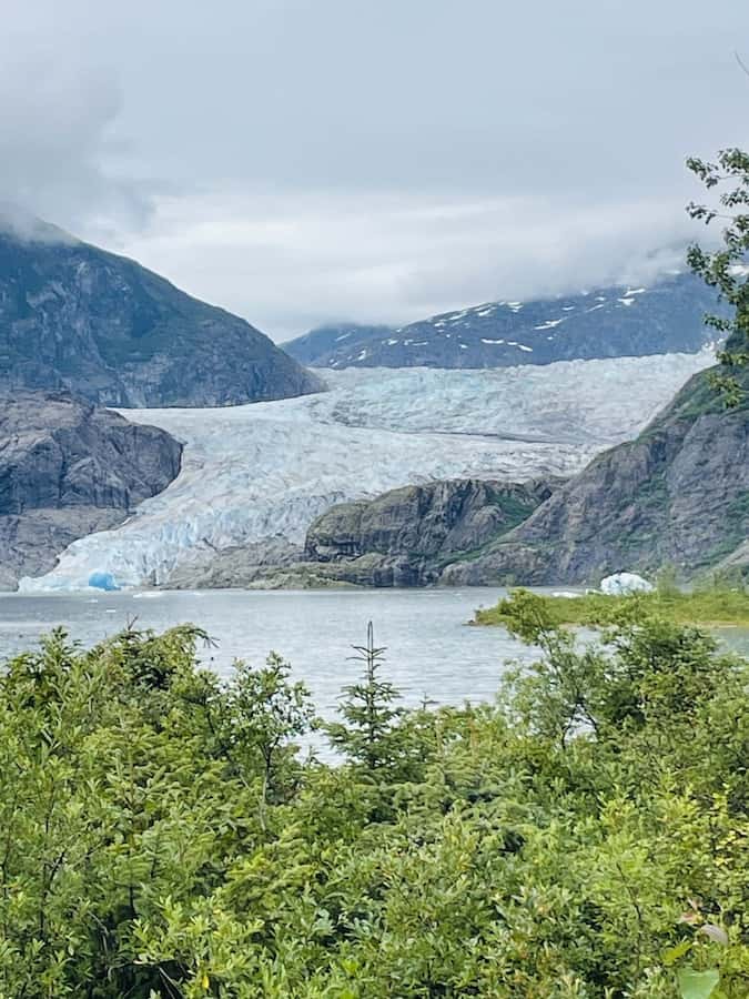 Alaskan scenery: glacier in the background with rock carved by the glacier towering around, blue water in the mid ground and green trees in the foreground