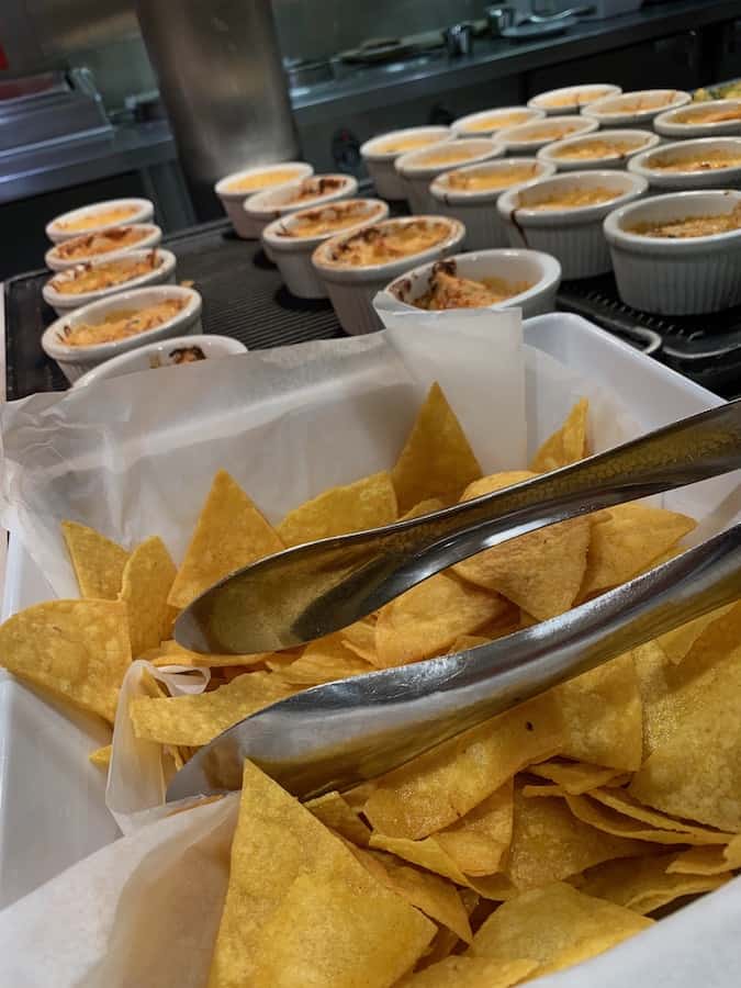 a buffet with a bowl of gluten-free tortilla chips with metal tongs, in the background rows of ramekins with artichoke dip
