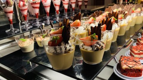 a dessert display with rows of strawberry parfaits in a long stemmed cup, two different types of custards in shorter cups, and plates of red velvet cake