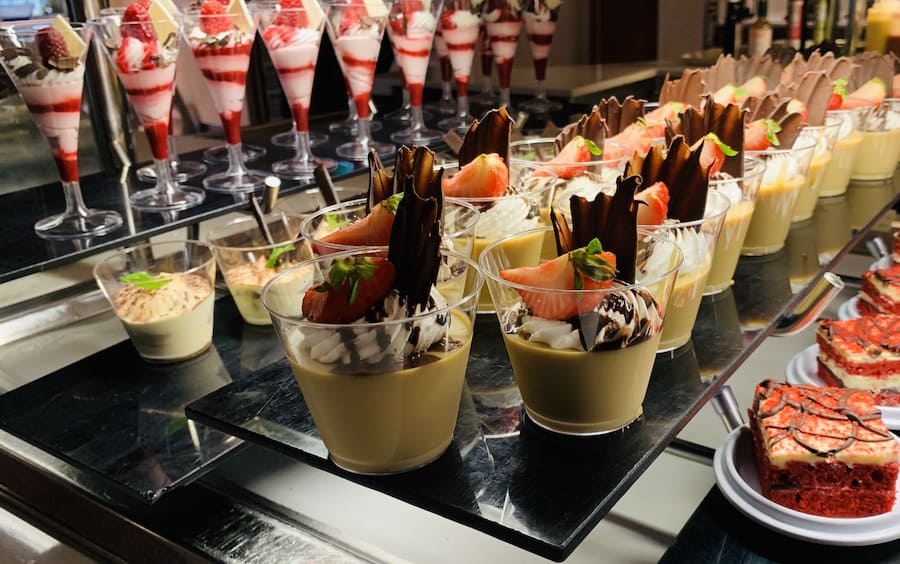 a dessert display with rows of strawberry parfaits in a long stemmed cup, two different types of custards in shorter cups, and plates of red velvet cake