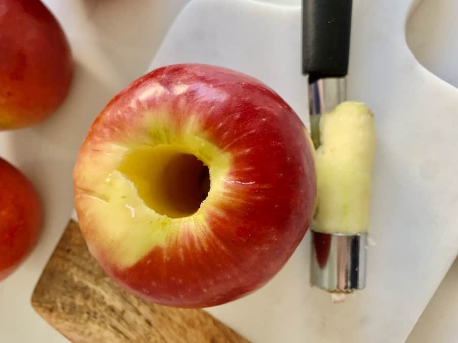 top view of a cored pink lady apple and an apple corer (with the core still in it) sitting on a marble and wood cutting board, whole pink/red apples in the background