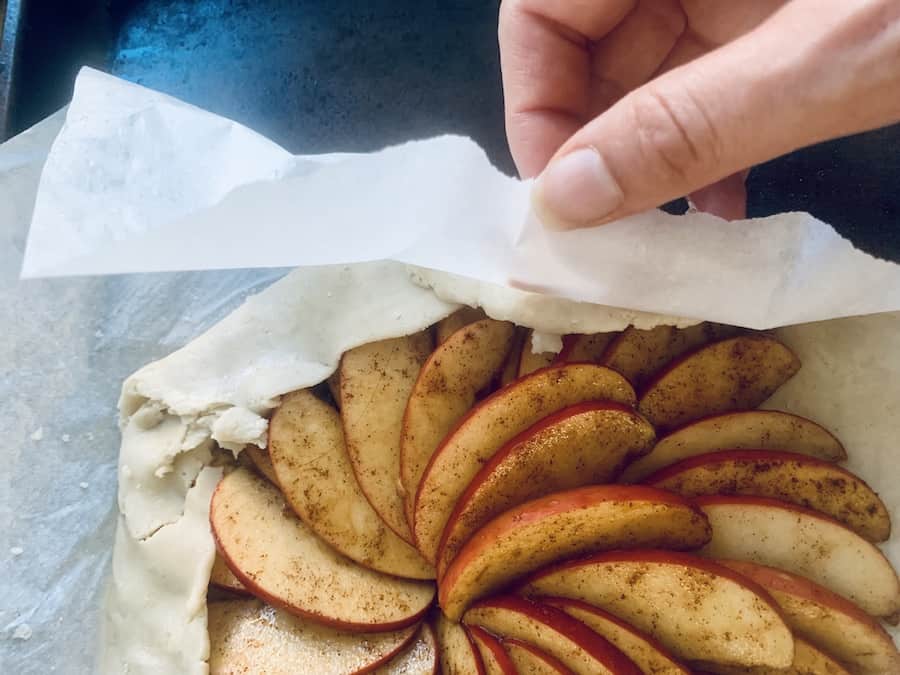 a hand pulling parchment paper off the edge of folded over pie crust dough for an apple galette