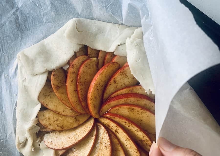 a hand pulling parchment paper off the edge of folded over pie crust dough for an apple galette, top view
