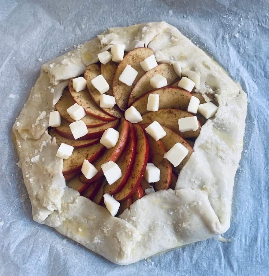 top view of raw apple galette with the dough folded over the apple filling and dots of butter scattered over the filling, and an egg wash and sprinkled sugar on the crust