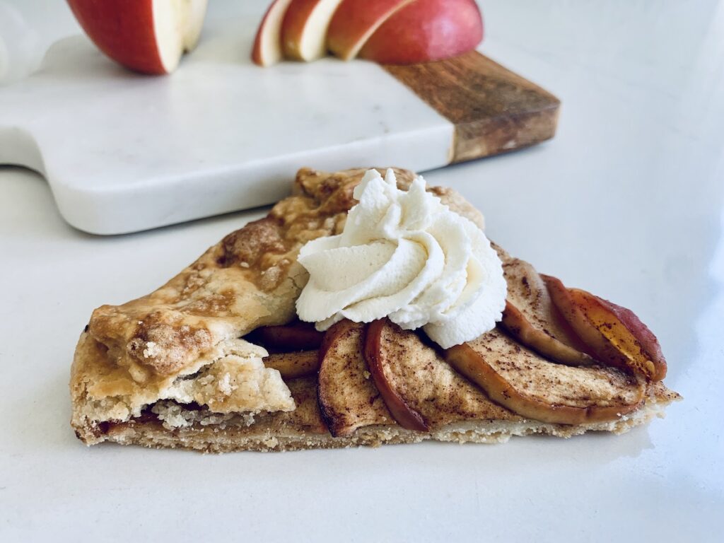 a slice of apple galette topped with whipped cream, apple slices and cutting board in the background