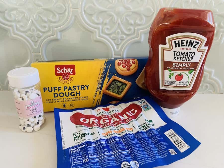 a bottle of candy eyes, a box of Schar Gluten-Free Puff Pastry, a package of AppleGate Organic Hot Dogs, and a bottle of Heinz Simply Ketchup, on a counter