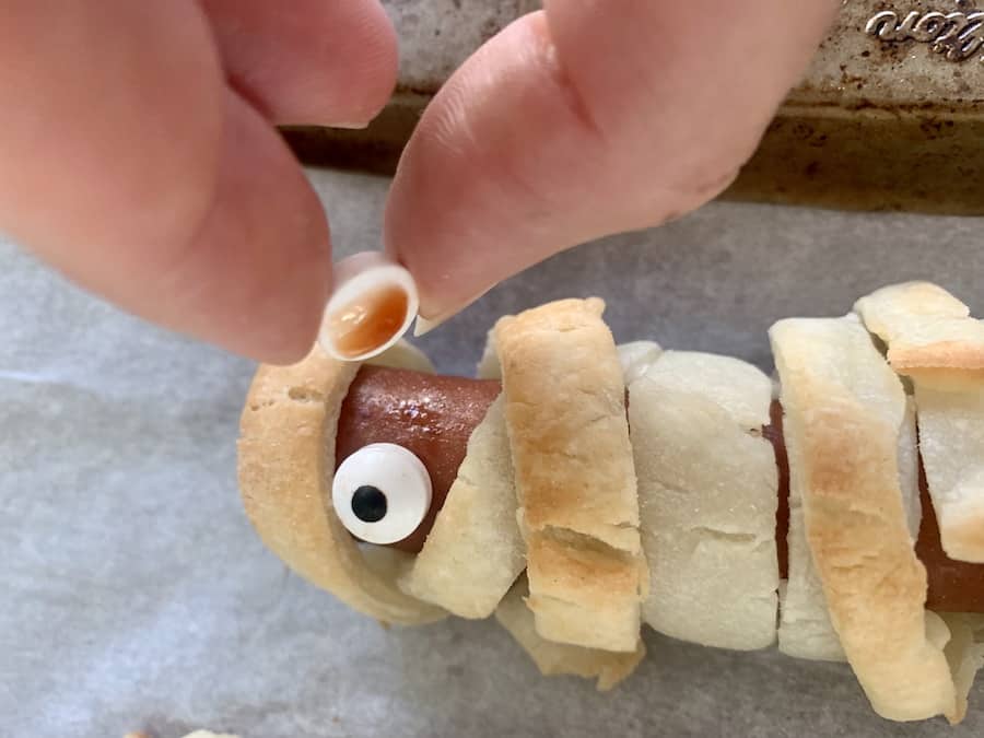 fingers placing a candy eye (with a dot of ketchup as glue) on a gluten-free mummy hot dog wrapped in gluten-free puff pastry with one candy eye