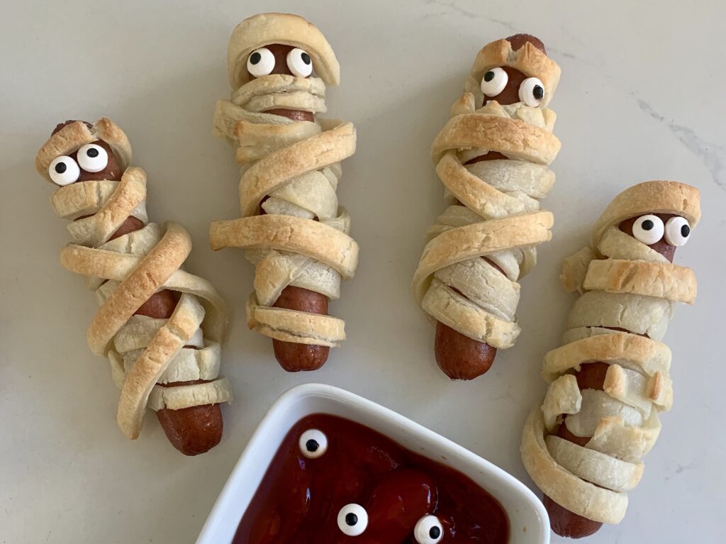 a gluten-free mummy hot dog wrapped in gluten-free puff pastry with candy eyes, a bowl of ketchup with candy eyes floating in it