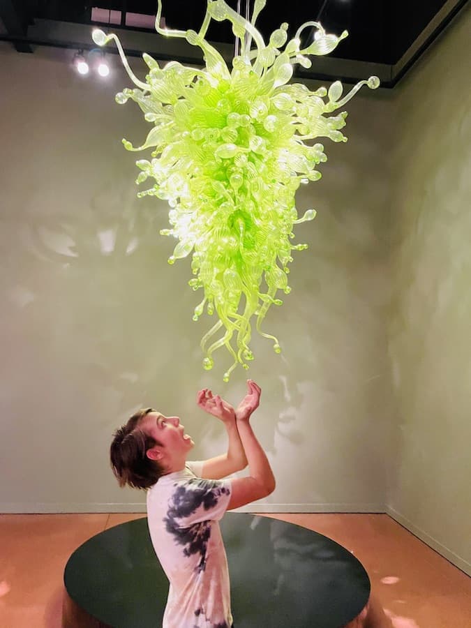 CJ posing for a photo under what appears to be an upside down Christmas tree made of bright green blown glass, he's holding his hands and looking surprised as if he were creating the spriraling green, glass object...