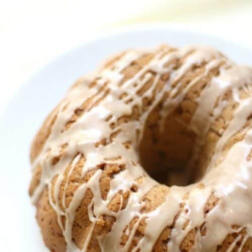 A small tan colored bundt cake, drizzled with white icing, on a white plate.