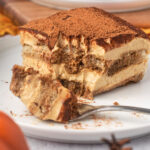 A slice of pumpkin tiramusu with visible light orange creamy layers and chocolate layers, with one bite on a fork.