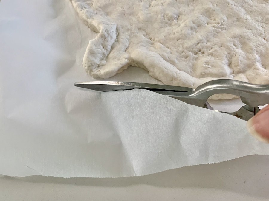 Scissors cutting the parchment paper around the dough, stem-portion of pumpkin-shaped pizza dough is visible.