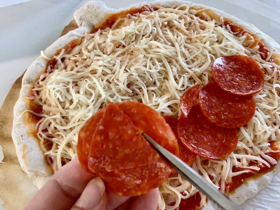 Scissors cutting triangle shapes out of pepporoni with gluten-free pumpkin pizza topped with sauce, cheese, and a pepperoni smile in the background.