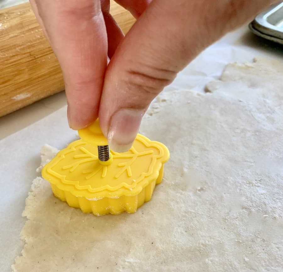 yellow leaf-shaped pastry cutter being used to cut out a leaf shape from rolled out pie dough, rolling pin in the background