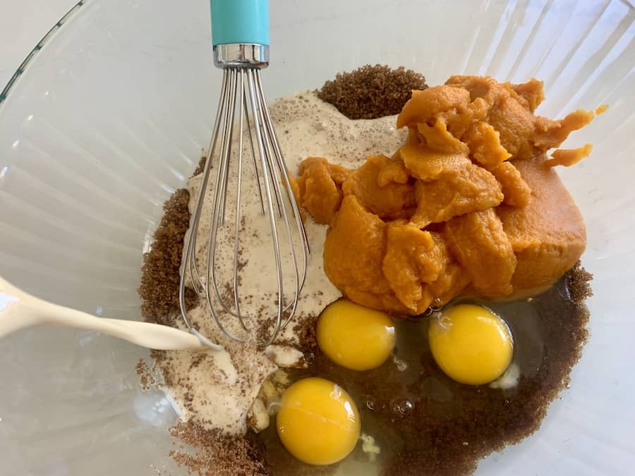 evaporated milk being poured into ingredients in a large glass mixing bowl, visible eggs, liquid, milk, pumpkin puree, brown sugar, and aqua whisk