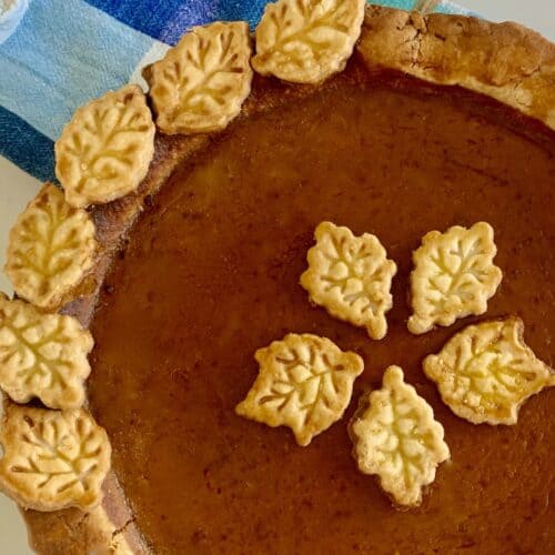 birds eye view: most of a pumpkin pie with pastry leaf shapes decorating the side and five in the center, plaid blue/aqua dish towel in the background