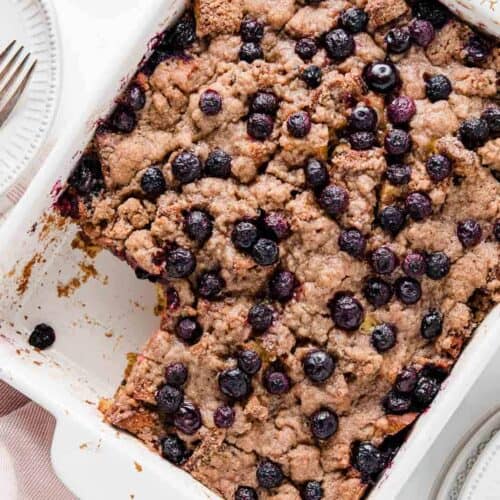 Gluten-Free baked French toast covered with blueberries, in a white casserole dish with a slice missing.