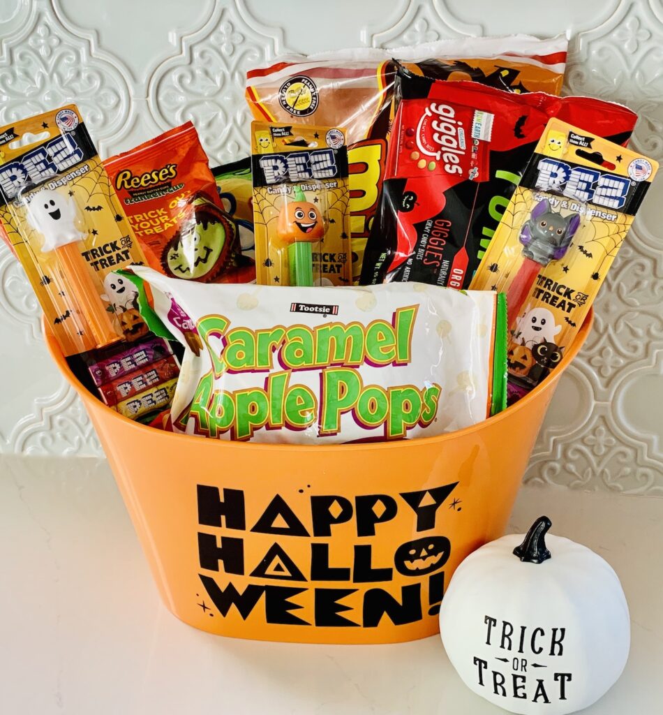 Orange bucket with words "Happy Halloween Halloween" full of packages of Halloween candy and a bag of "Caramel Apple Pops" visible in the front of the bucket. Next to the bucket is a white pumpkin with black words: "Trick or Treat".