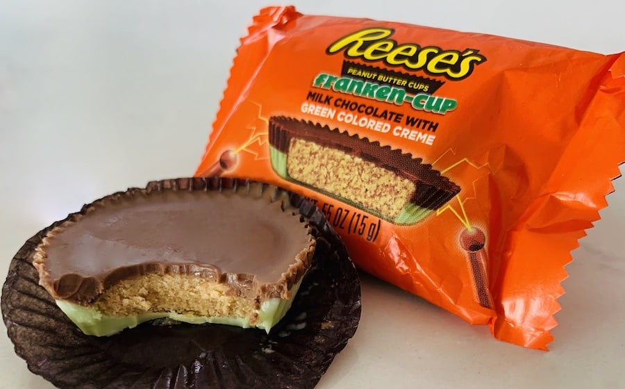 Reese's Peanut Butter "Franken-Cup" with green-colored white chocolate on the bottom and a bite missing. A package of a single Reese's Franken-cup in the background.