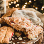 Gluten-Free cranberry & orange scones on wooden board with icing drizzled on top of the scones and a little bit on the board. Twinkling lights on a tree in the background.