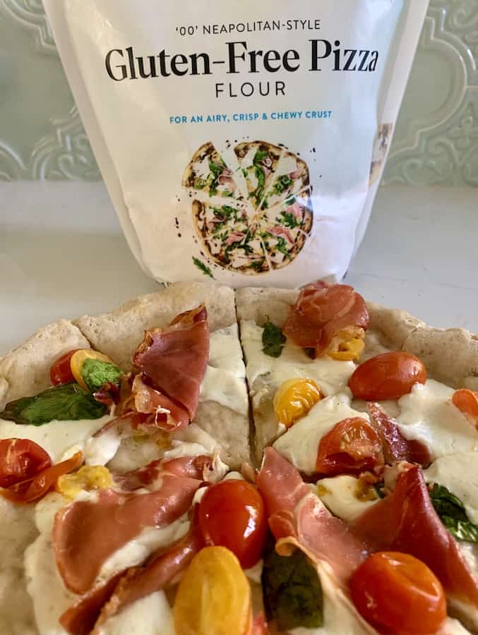 part of a gluten-free Neopolitan pizza topped with cheese, tomato, basil, and prosciutto. Bag of 00 Neopolitan Style Gluten-Free Pizza Flour in the background.