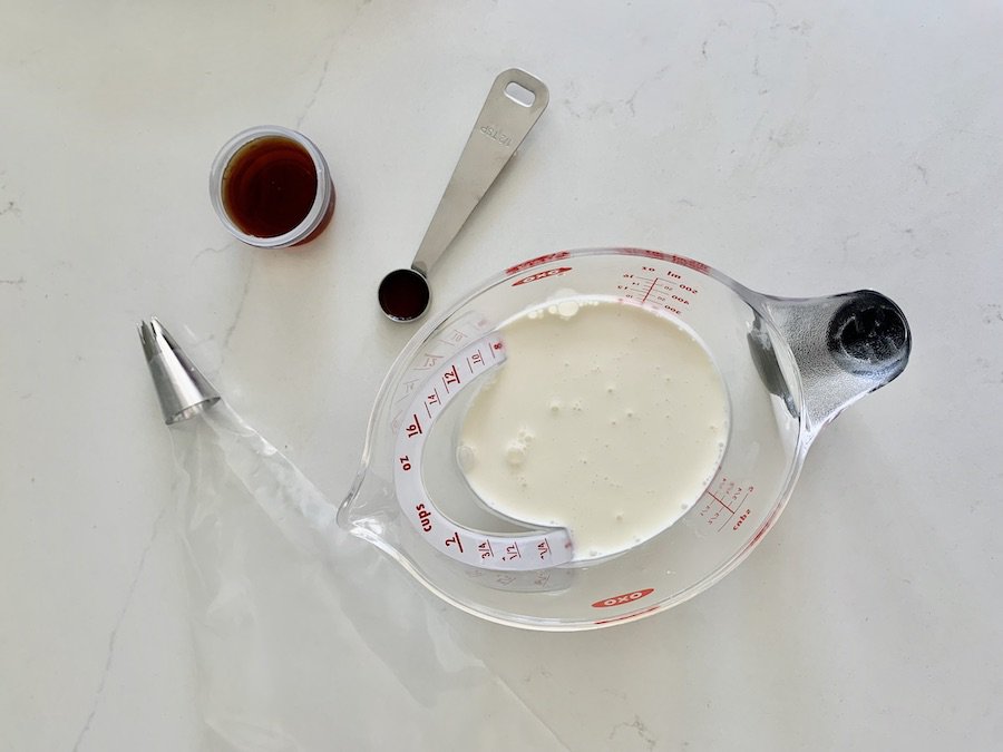 birds eye view of measuring cup of heavy cream, measuring spoon of vanilla, small cup of maple syrup and empty piping bag with star tip