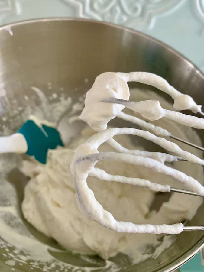 finished whipped cream on two beaters, in the background: a metal mixing bowl with whipped cream and an aqua spatula