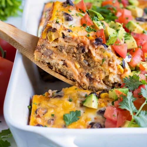 Layered Mexican casserole with cheese, meat, tortillas, and topped with tomatoes and cilantro. One slice being lifted out of a white casserole dish by a wooden spoon.