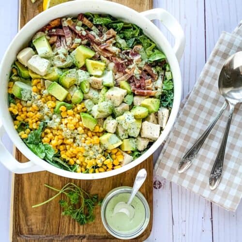 Birds eye view of a bowl of green goddess Cobb salad, with utensils on a gingham napkin and a small bowl of creamy-green dressing.