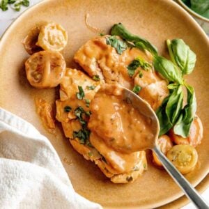 Spoon pouring creamy tomato sauce on chicken breasts on a plate.