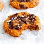 A bright orange sweet potato oat cookie with chocolate drizzle on top and bite taken out. More cookies in the background.