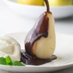 A pear standing up, half coated with chocolate sauce, on a white plate with whipped cream and mint leaves on the side, and a bowl of pears in the background.