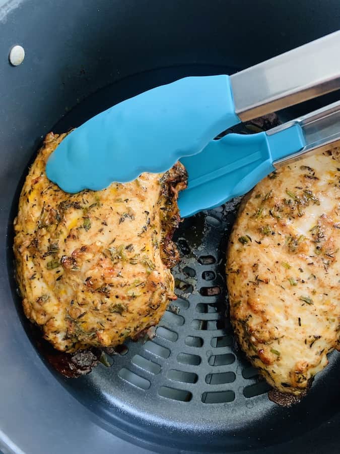 Air fryer basket containing two partially cooked turkey tenderloin breasts coated with marinade and spices. Aqua tongs rotating one of the turkey tenderloins.