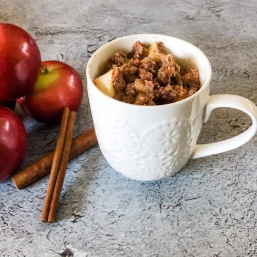 Apple crumble in a white mug on a grey counter, with cinnamon sticks and apples next to it.
