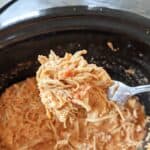 Spoon holding shredded chicken over a crock pot of more shredded chicken in a creamy salsa sauce.