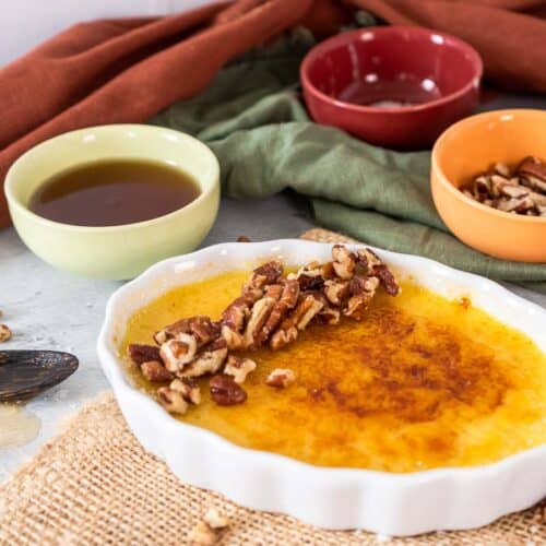 Creme brûlée topped with pecans, in a white ramekin. Small bowls with dark liquid and nuts in the background along with fall colored napkins.