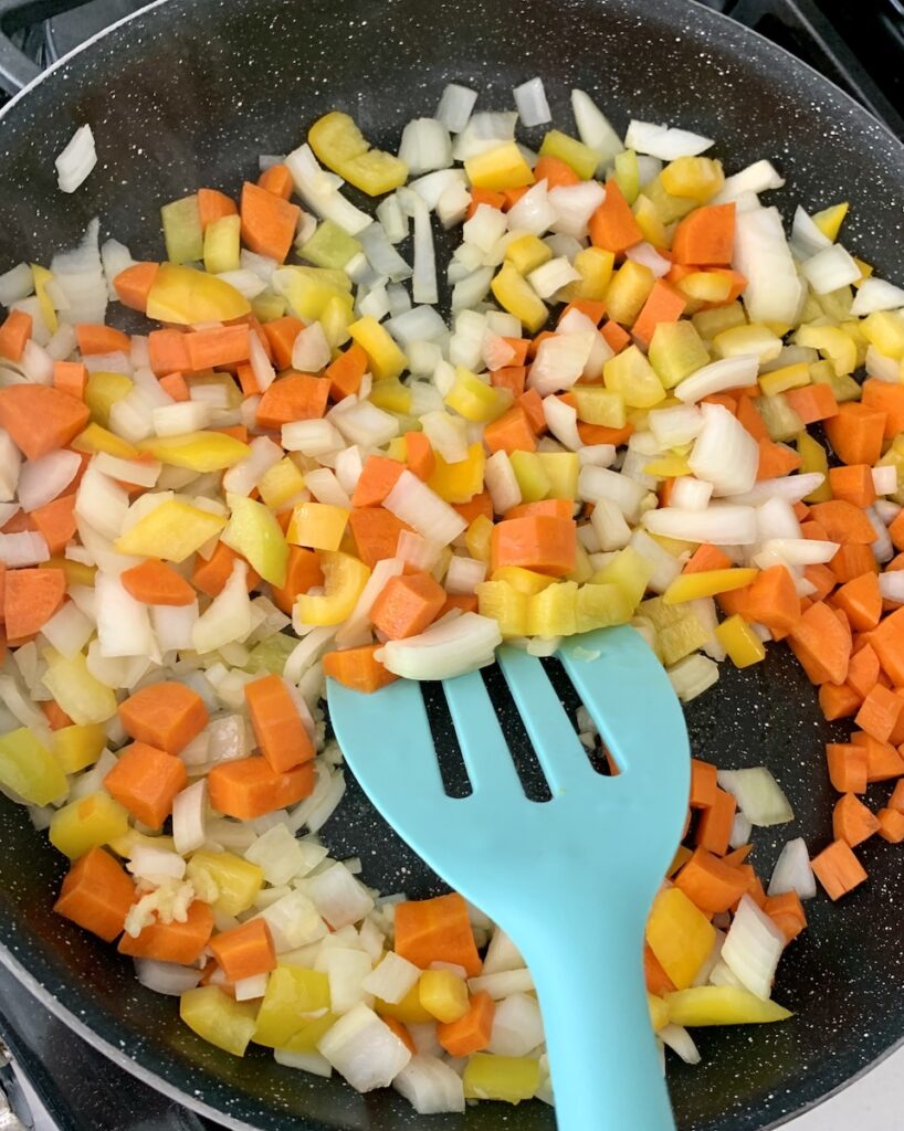 An aqua spatula stirring carrots, peppers, onions, and garlic in a frying pan.