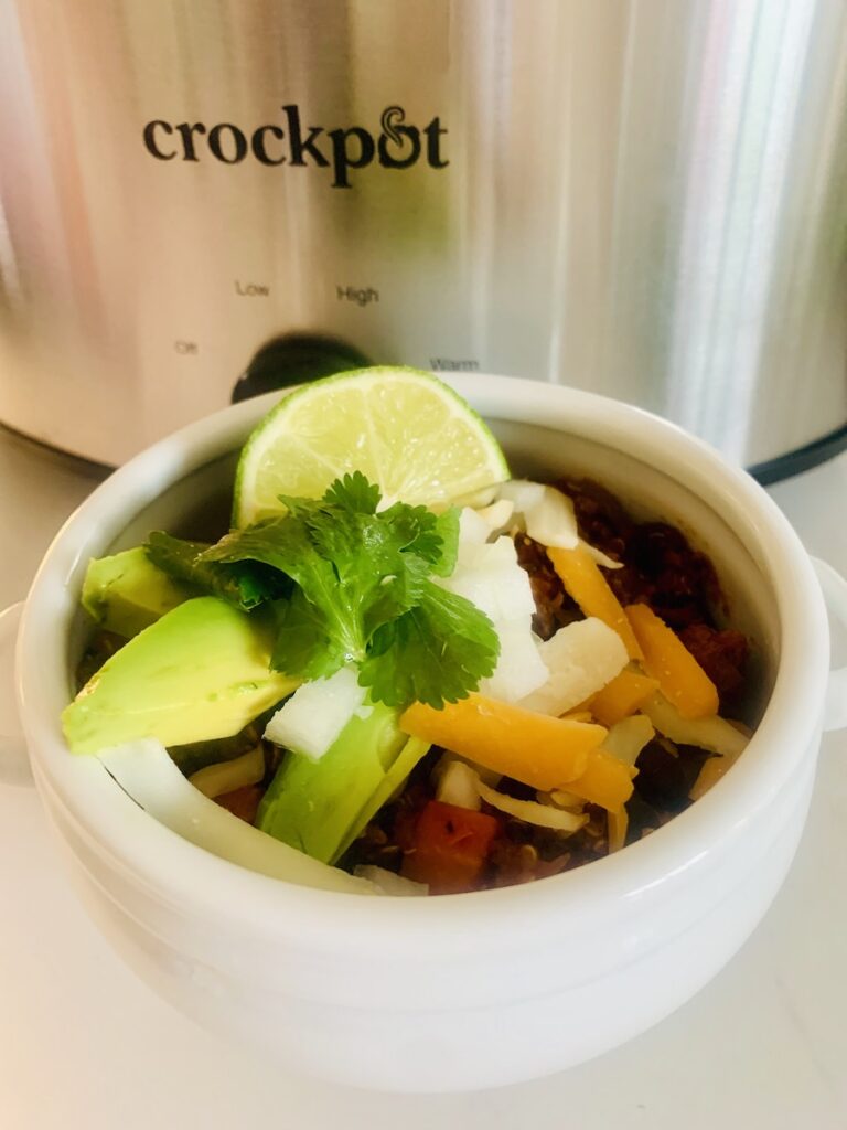 Chili in a white bowl, topped with avocado, lime, cilantro and cheese. Part of a crockpot in the background.