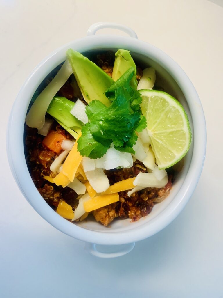 Chili in a white bowl, topped with avocado, lime, cilantro and cheese.