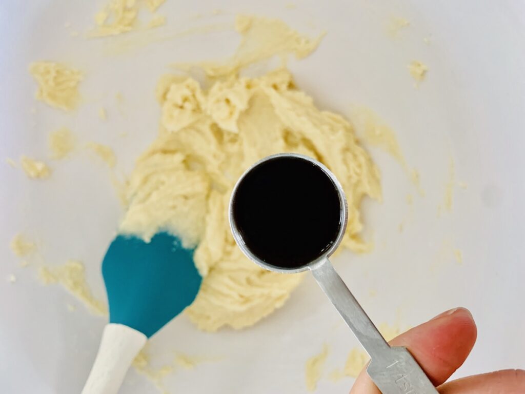 A teal spatula scraping the batter in the bowl. A teaspoon of vanilla in focus above the bowl of batter.