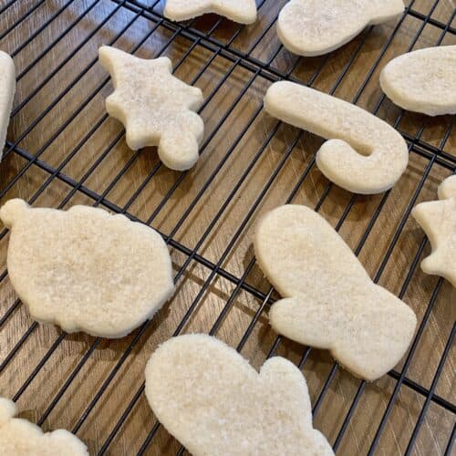 Unfrosted, baked, Gluten-Free sugar cookie cut outs on a cooling rack. Shapes include mittens, holly, Santa, and a candy cane.