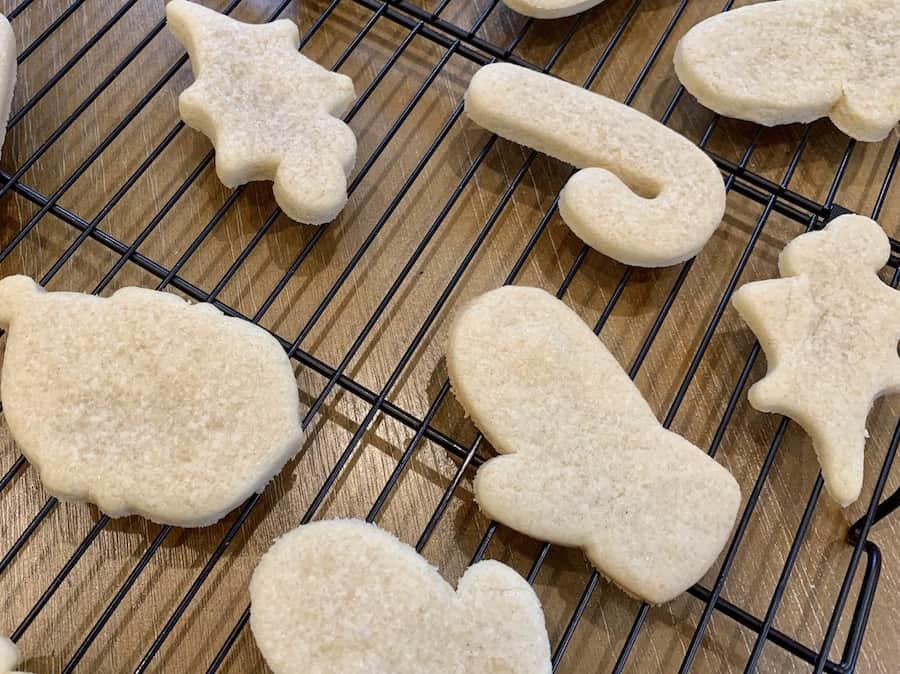 Unfrosted, baked, Gluten-Free sugar cookie cut outs on a cooling rack. Shapes include mittens, holly, Santa, and a candy cane.