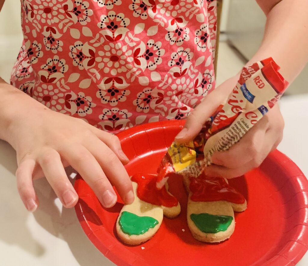 Child's hands using store-bought red icing to decorate mitten-shaped cookies, that already have green frosting piped on.