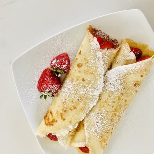 Birds eye view: 2 crepes filled with strawberries and topped with powdered sugar on a square white plate with two strawberries.