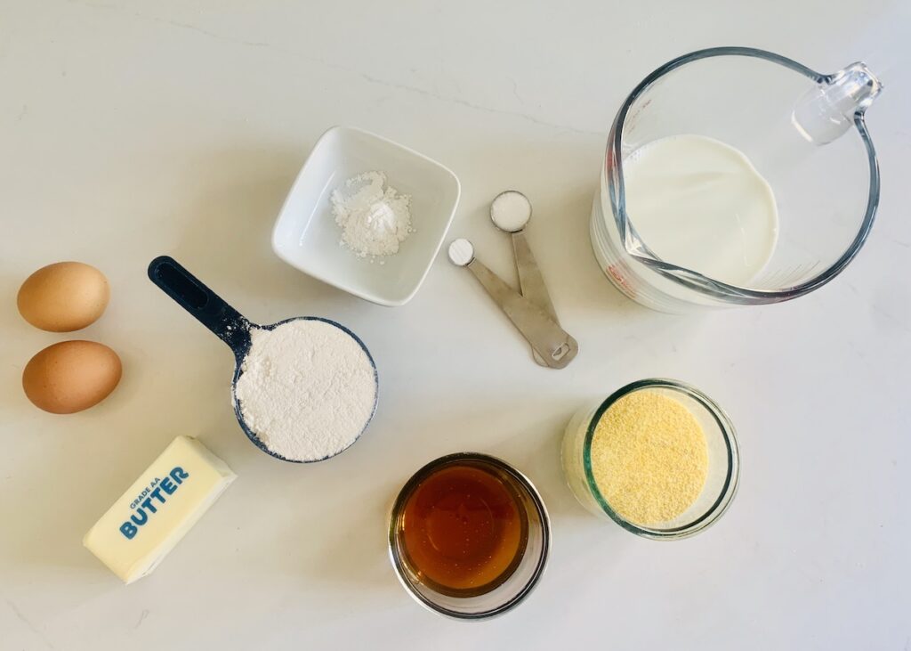 Birds eye view: ingredients on a counter: butter stick, measuring cup with flour, measuring spoons and small square white bowl with white ingredients, glass cups with golden honey and yellow cornmeal, andmeasuring cup with milk.