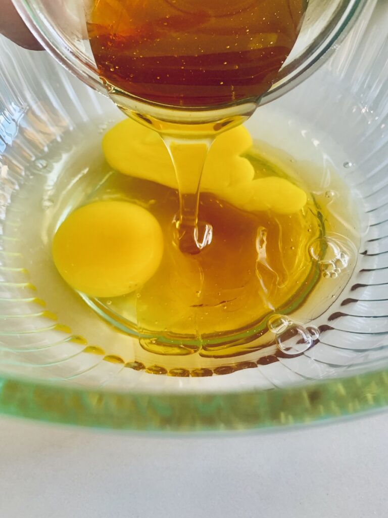 Golden honey being poured into a glass bowl with two eggs.