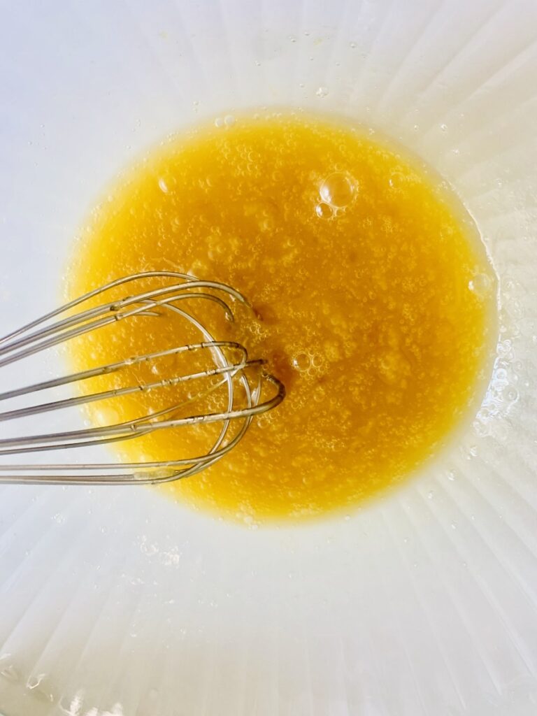 Whisk resting in a bowl of mixed eggs and honey, golden colored liquid.