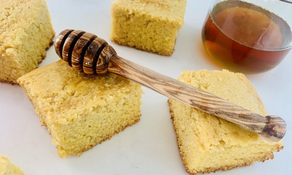 Squares of gluten-free honey cornbread scattered on a white counter top. Honey in a glass cup on the counter. A honey dipper resting on two pieces of cornbread.