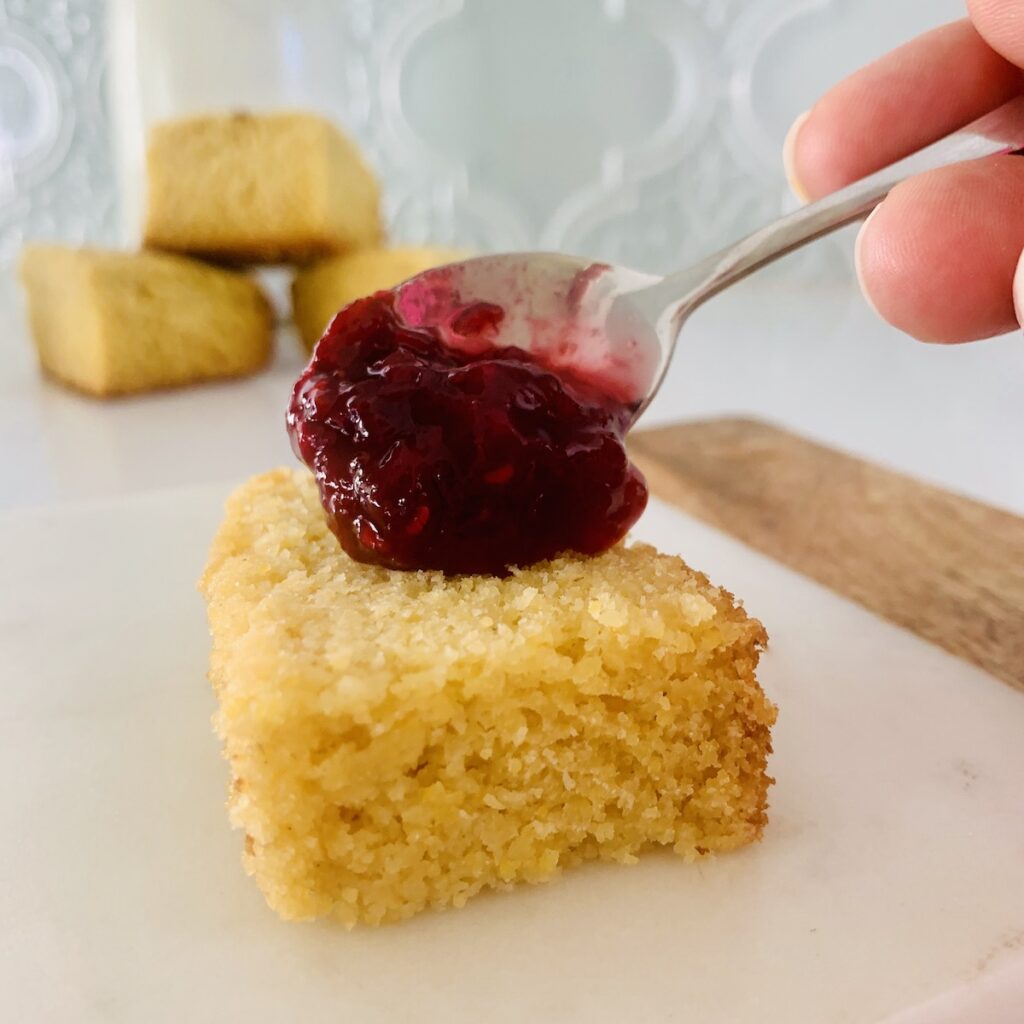 Small spoon scooping red jam onto a cornbread square. Cornbread and milk in the background.