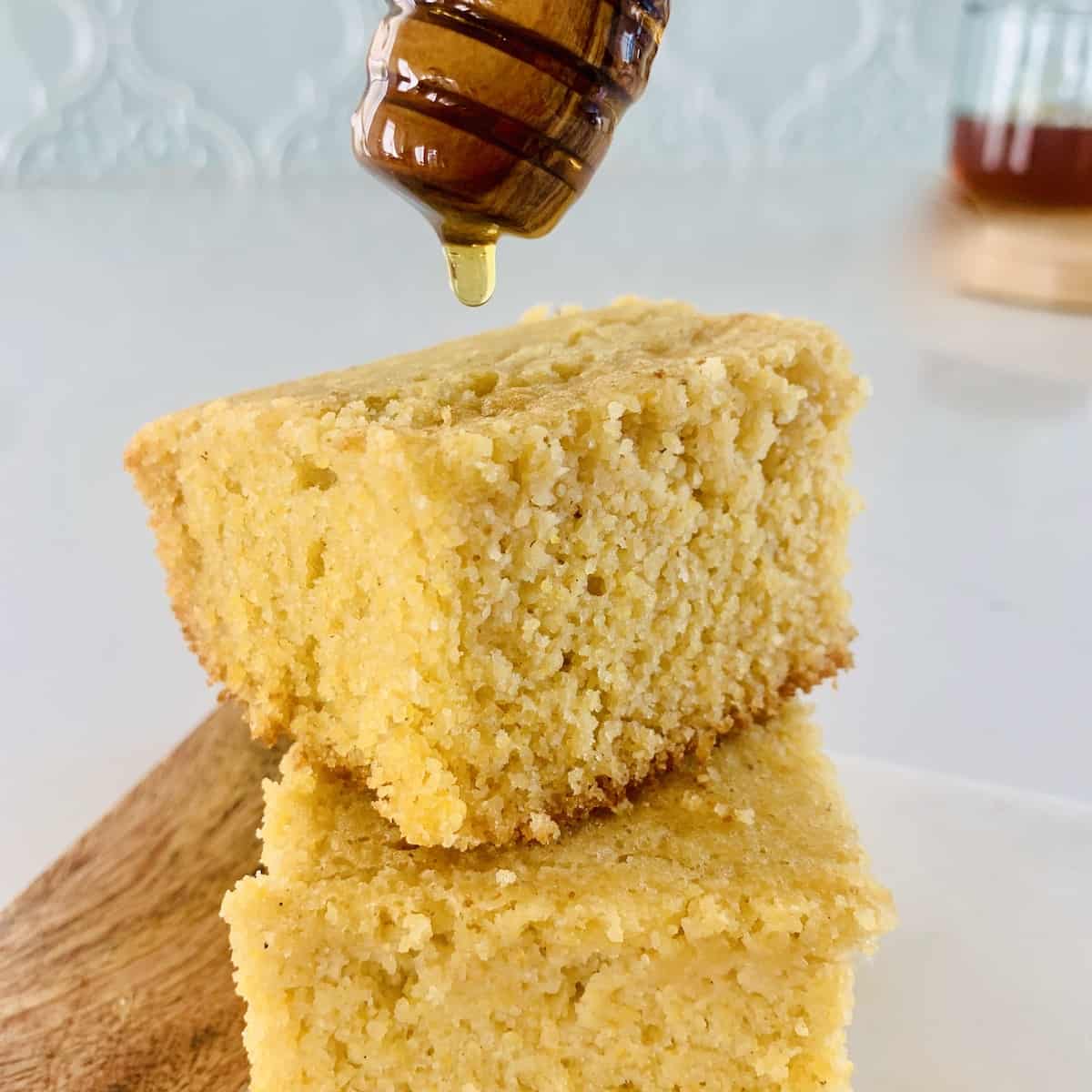 2 squares of gluten-free cornbread stacked, with honey being poured on top by a honey dipper.